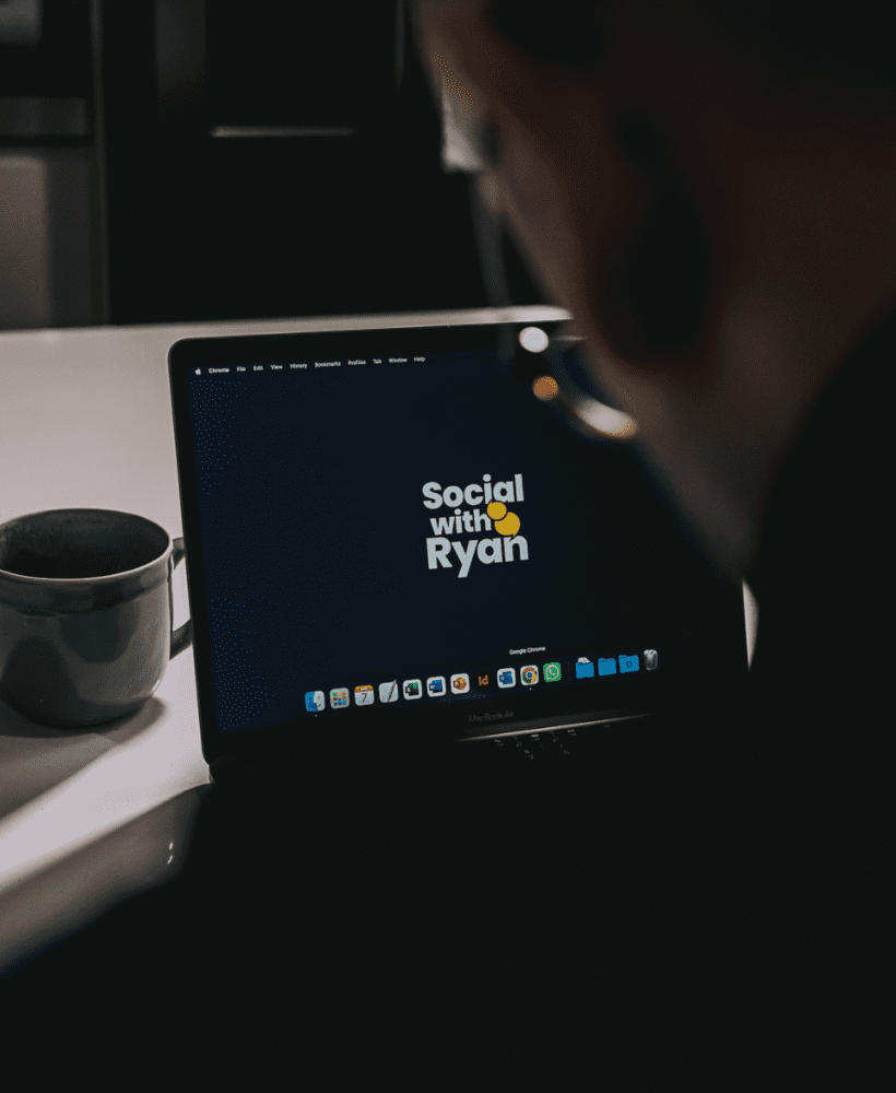 The picture is taken over a man's shoulder, with the man working on a laptop. The laptop has the words 'Social with Ryan' on the screen. A mug sits next to the laptop.