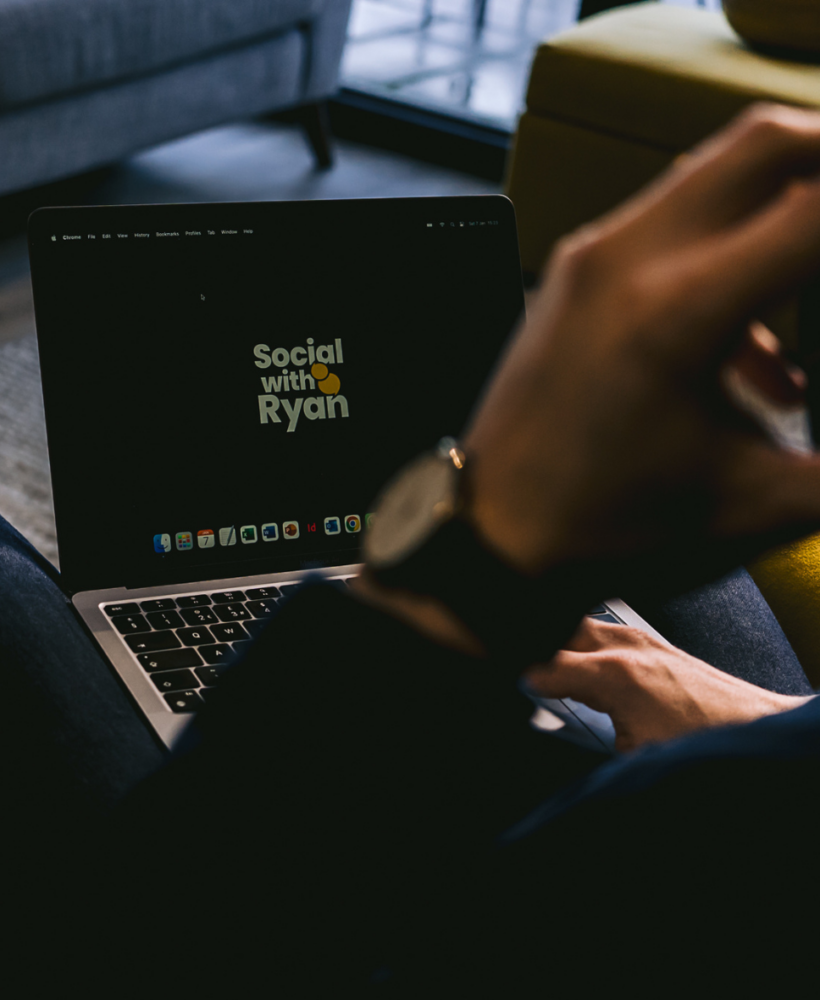 An over-the-shoulder shot of man sat with a mobile phone to his ear and browsing the 'Social with Ryan' site on his laptop