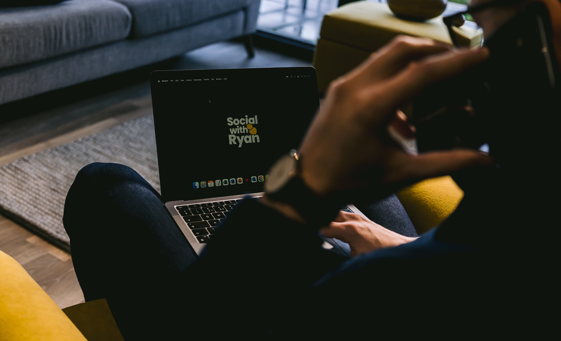 An over-the-shoulder shot of man sat with a mobile phone to his ear and browsing the 'Social with Ryan' site on his laptop